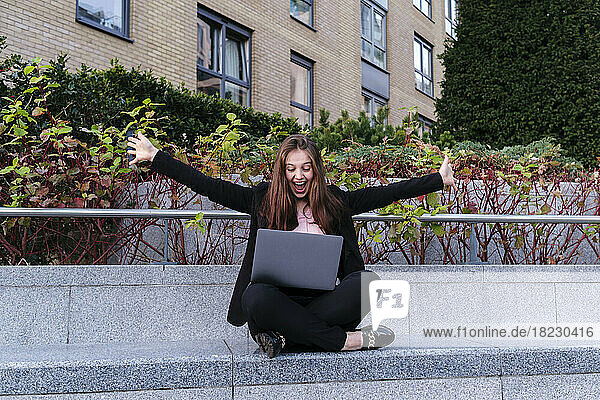 Happy businesswoman with arms outstretched with laptop sitting on bench