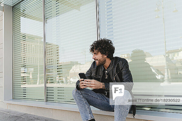 Lächelnder Mann mit Mobiltelefon sitzt auf der Fensterbank