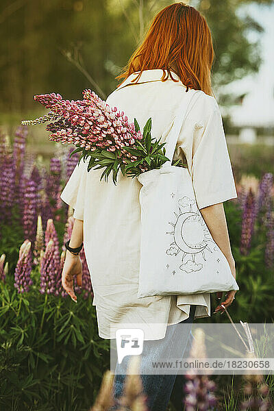 Redhead girl walking with lupin flowers in tote bag on field