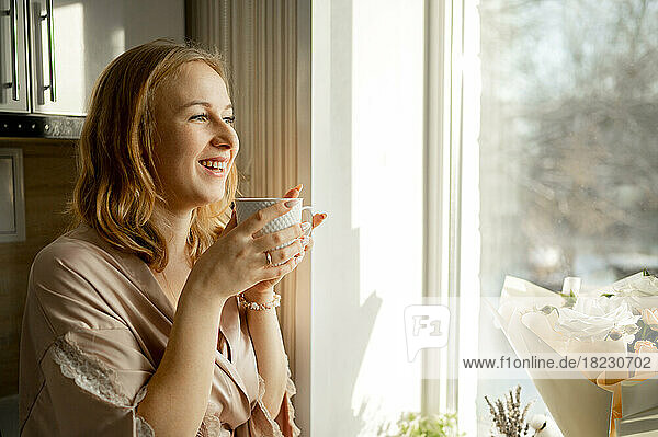 Happy young woman with coffee cup looking out through window