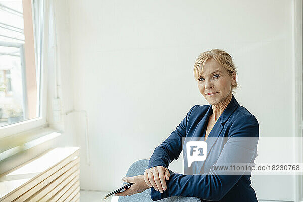 Businesswoman with smart phone leaning on armchair in front of white wall