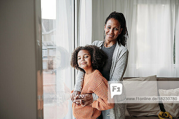 Happy mother and daughter standing together by window at home