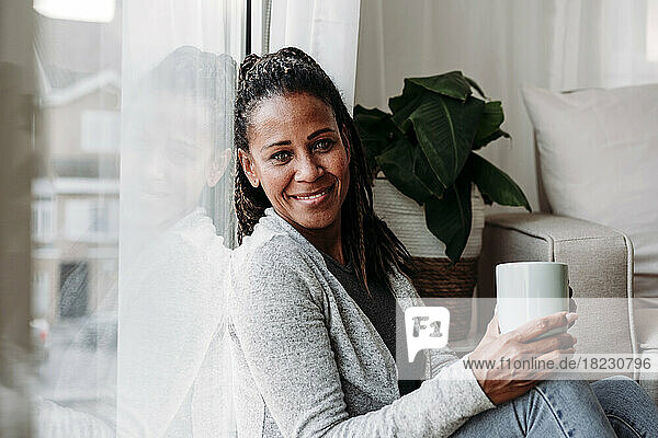Lächelnde reife Frau mit Kaffeetasse lehnt zu Hause an Glasfenster