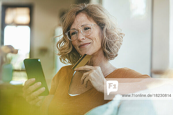 Lächelnde reife Frau mit Kreditkarte und Smartphone zu Hause