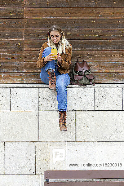 Young woman using smart phone sitting on wall