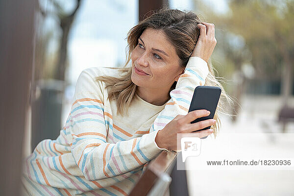 Thoughtful young blond woman sitting on bench with smart phone
