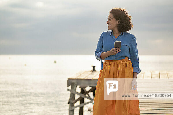 Smiling woman standing with mobile phone on jetty at sunset