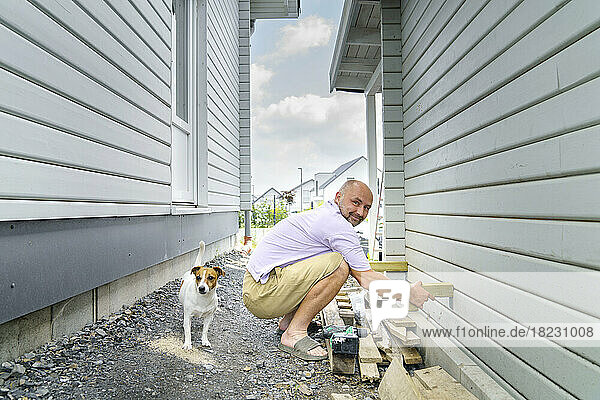 Smiling man building wooden wall crouching by dog near house