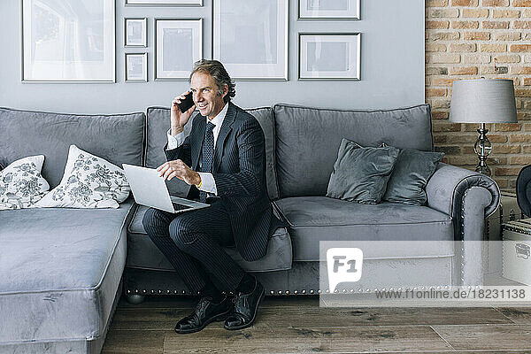 Smiling mature businessman talking through mobile phone sitting on sofa at home