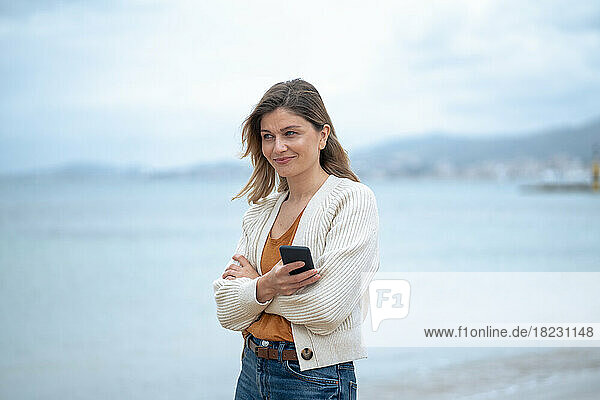 Smiling beautiful young woman with arms crossed holding smart phone at beach