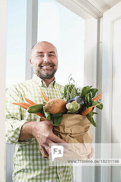 Smiling man holding fresh organic vegetables in paper bag near doorway
