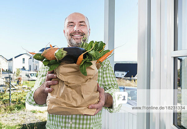 Smiling man holding paper bag full of vegetables at doorway