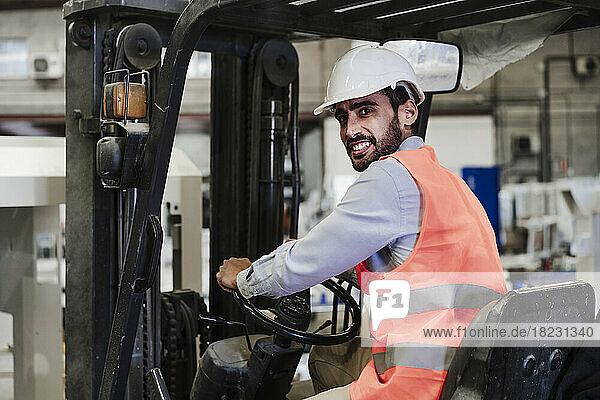 Happy engineer wearing protective workwear driving forklift in industry