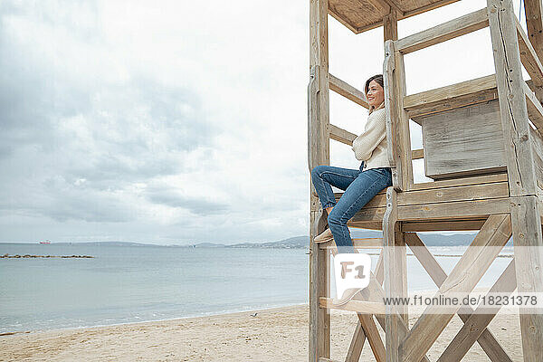 Smiling young woman sitting on wooden lifeguard hut at beach