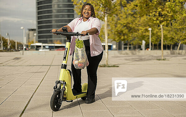 Happy curvy woman with electric push scooter and bag of vegetables standing at footpath