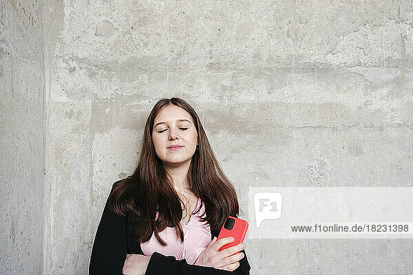 Young woman with eyes closed holding mobile phone leaning on wall