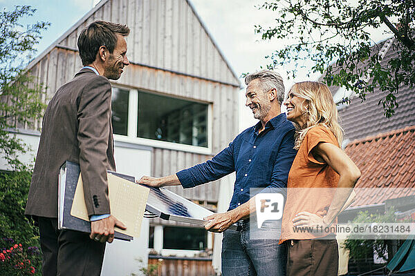 Happy real estate agent with couple holding solar panel in front of house