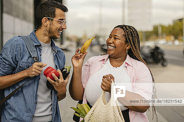 Cheerful young man with girlfriend holding bag of vegetables at footpath
