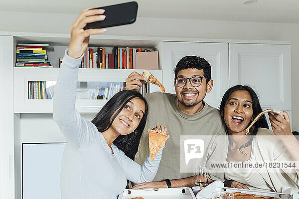 Smiling woman taking selfie through smart phone with woman and man showing pizza slice at home