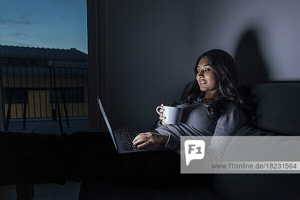 Smiling young woman holding coffee cup using laptop at home