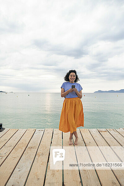 Lächelnde Frau steht mit Smartphone am Steg vor dem Meer