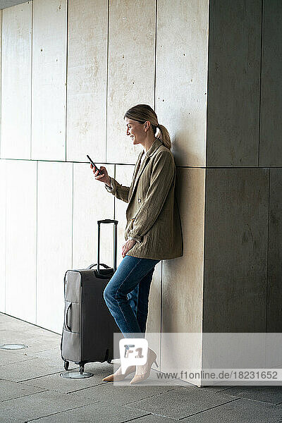 Smiling businesswoman using smart phone by luggage leaning on wall in tunnel
