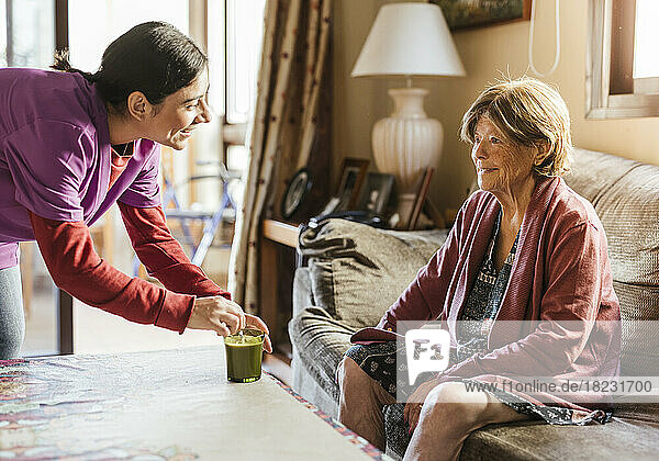 Happy caregiver stirring coffee and talking to senior woman sitting on sofa at home