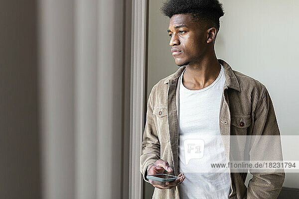 Thoughtful young man with smart phone looking out of window at home