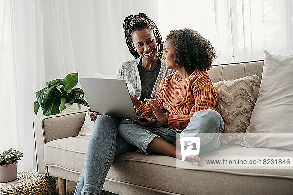 Happy mother and daughter sitting with laptop on sofa at home