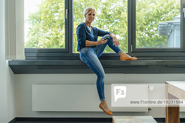 Businesswoman with mobile phone sitting on window sill at office