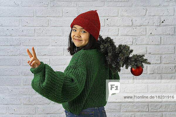 Smiling girl with Christmas tree gesturing peace sign