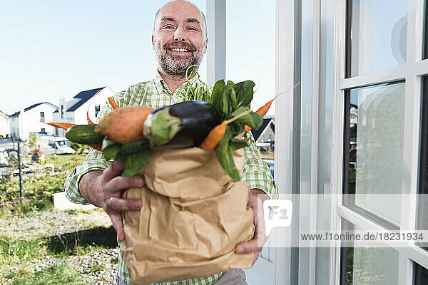 Smiling man holding bag full of fresh vegetables standing at doorway