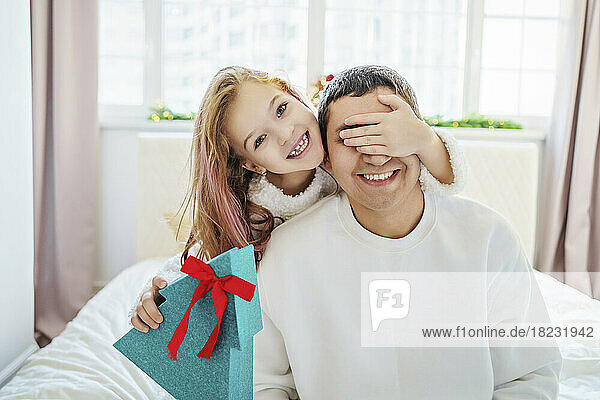Happy girl holding Christmas gift and covering eyes of father sitting on bed at home