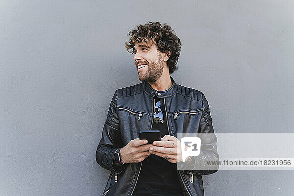 Glücklicher Mann in schwarzer Lederjacke hält Mobiltelefon vor grauer Wand