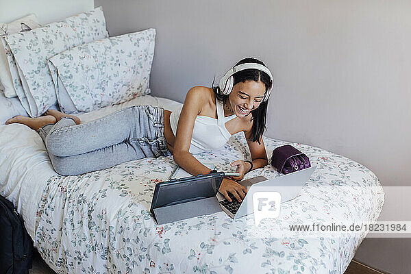 Glückliches Mädchen mit Kopfhörern  das zu Hause auf dem Bett liegt und drahtlose Geräte nutzt
