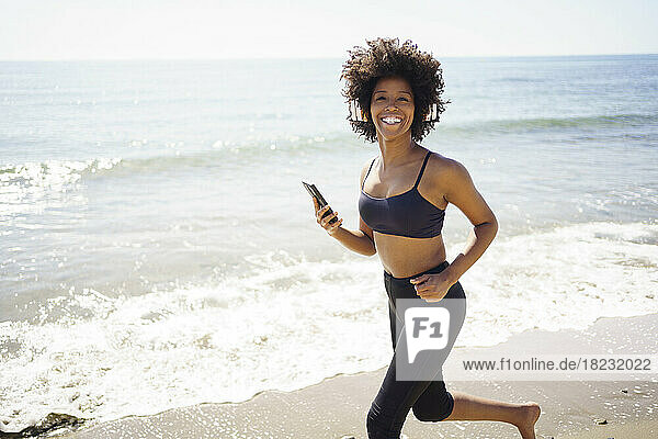 Happy young woman running with smart phone on shore at beach