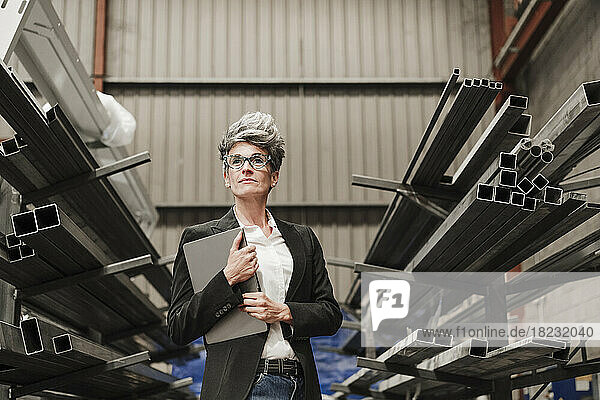 Mature businesswoman with laptop standing amidst metal beams on racks