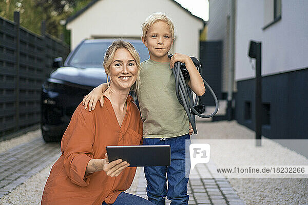 Mother with tablet PC by son holding electric vehicle charger plug in front yard