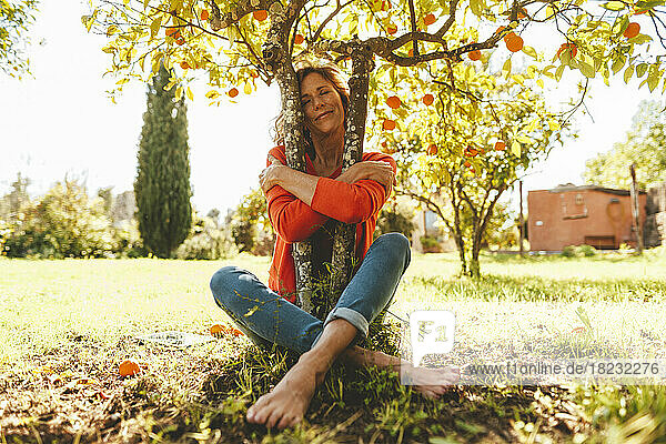 Reife Frau mit geschlossenen Augen umarmt einen Orangenfruchtbaum