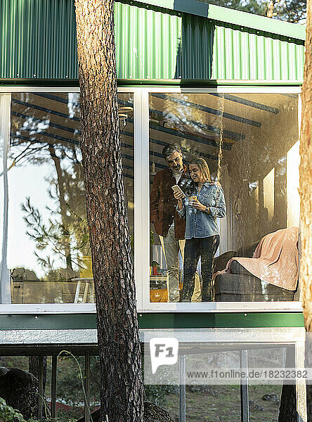 Älteres Paar steht am Fenster eines Hauses und schaut auf sein Mobiltelefon