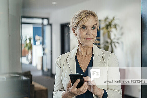 Thoughtful businesswoman with smart phone in office seen through glass