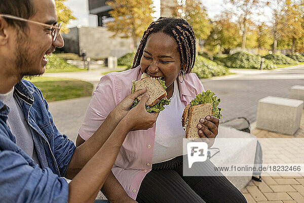 Glücklicher Mann teilt Sandwich mit Frau  die auf einem Sitz am Fußweg sitzt