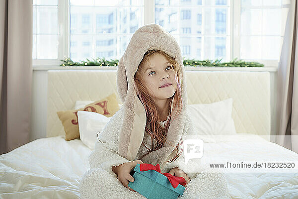 Girl wearing hat sitting with Christmas gift on bed contemplating at home