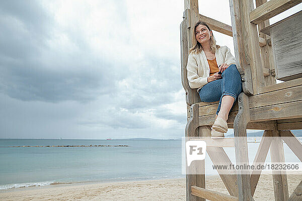 Lächelnde junge Frau sitzt auf einer hölzernen Rettungsschwimmerhütte am Strand unter bewölktem Himmel