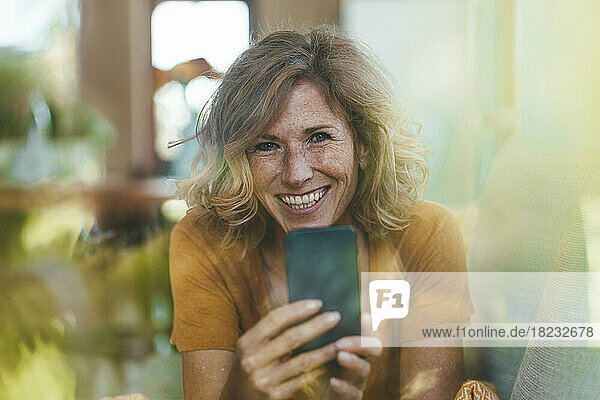 Glückliche Frau mit Smartphone  gesehen durch das Glas ihres Hauses
