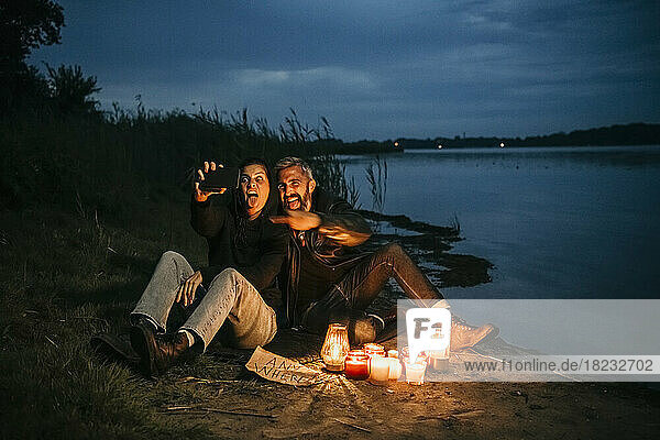 Glückliche Frau macht ein Selfie mit einem Mann  der bei brennenden Kerzen am Seeufer sitzt