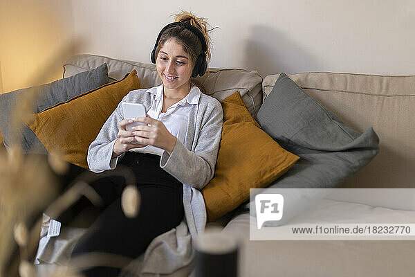 Happy young woman wearing wireless headphones using smart phone on sofa at home