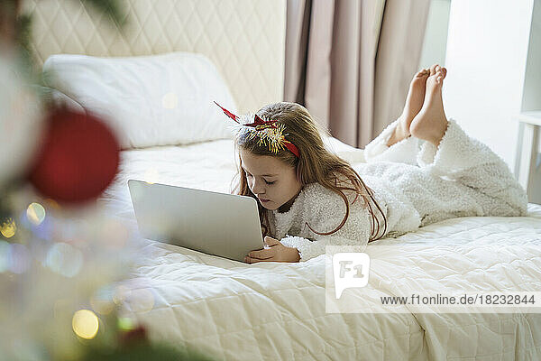 Girl wearing Christmas headband lying on bed and using laptop at home