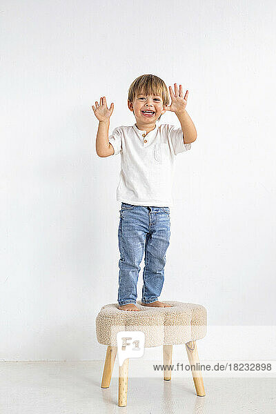 Smiling boy gesturing on stool in front of wall at home
