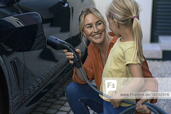Smiling woman holding electric vehicle charger plug looking at daughter in front yard